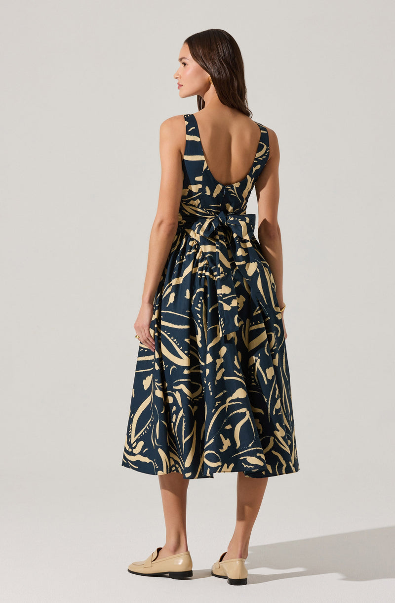 A woman is wearing a sleeveless, ankle-length dress with a deep scoop back. The dress features an abstract, bold navy blue and beige pattern, with a waistband tied into a large bow at the back. She is also wearing beige flat shoes.