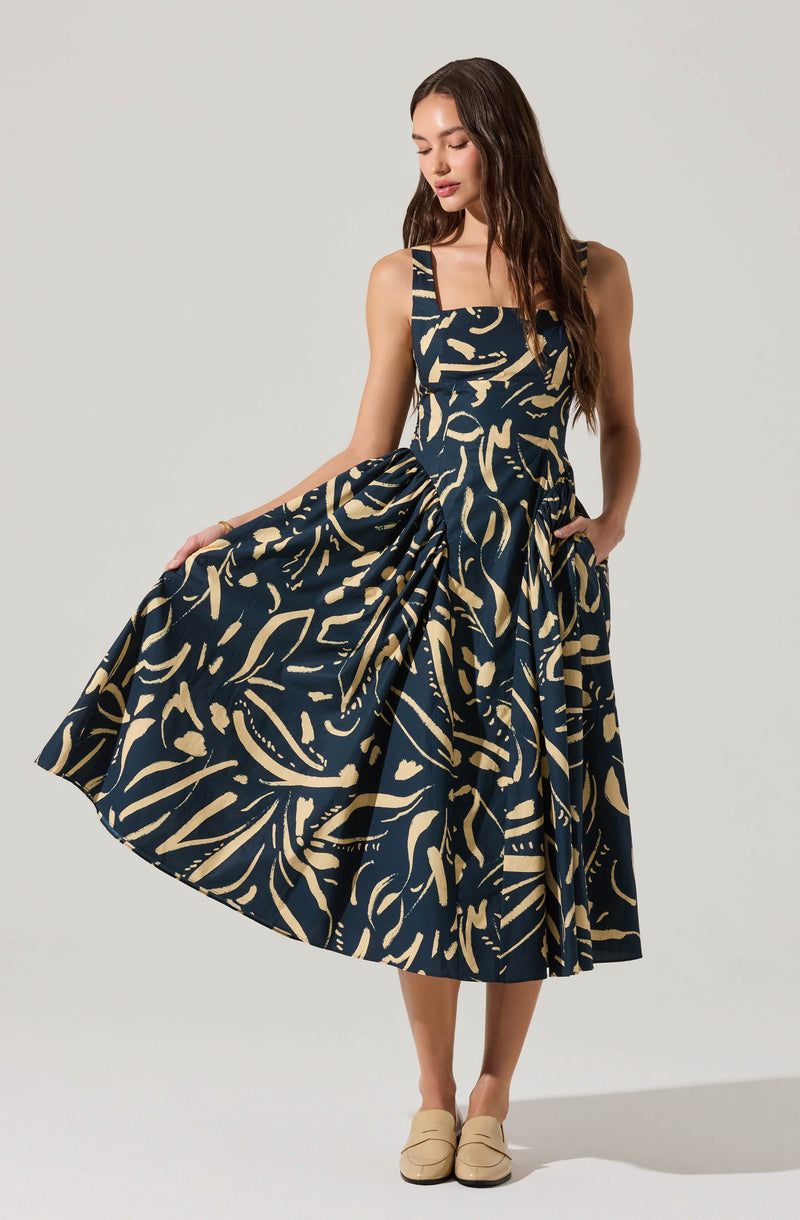 A woman is wearing a sleeveless, midi-length dress with wide straps. The dress features a dark background with an abstract beige pattern throughout. She is holding the side of her skirt with one hand, slightly lifting it, and has her other hand in a pocket. She is also wearing beige slip-on shoes.