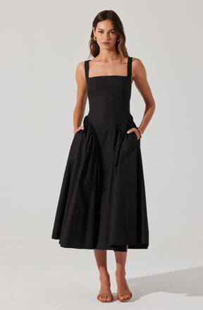 A woman is wearing a sleeveless black dress with wide straps and a fitted bodice. The dress has a full, gathered skirt that extends to mid-calf length. She has her hands in the pockets of the dress and is accessorizing with gold hoop earrings and a bracelet. She is wearing nude-colored, open-toe high heel shoes.
