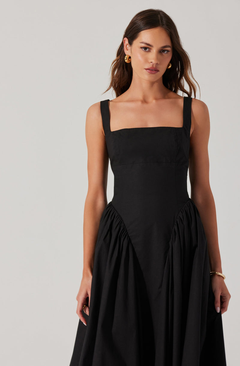 A woman is wearing a black sleeveless dress with wide shoulder straps. The bodice is fitted and square-necked, extending into a gathered skirt that falls to the floor. She accessorizes with gold hoop earrings and a gold bracelet on her wrist.