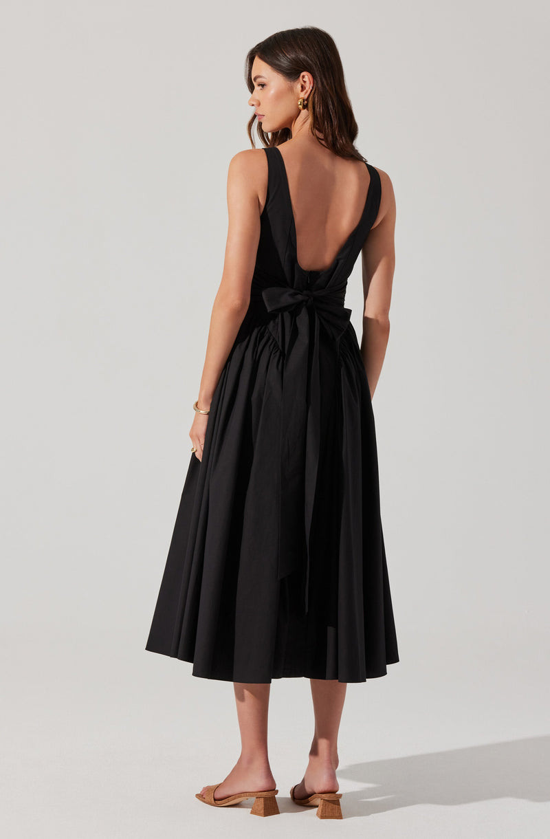 A woman is wearing a sleeveless, black midi dress with a deep V-back and a waist tie bow. The dress has a full, gathered skirt that falls below the knees. She is accessorized with gold hoop earrings and a gold bracelet, and is wearing tan open-toe sandals with a block heel.