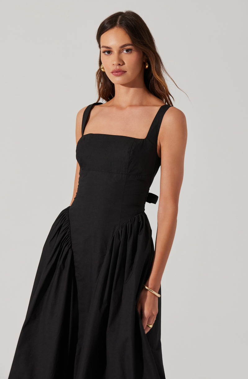 A woman is wearing a sleeveless black dress with wide shoulder straps. The dress has a fitted bodice and a gathered, full skirt. She accessorizes with gold hoop earrings, a gold bangle on her wrist, and a ring.