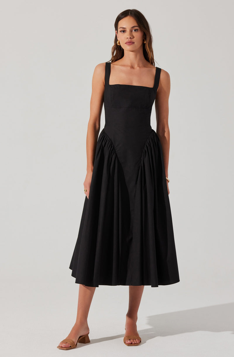 A woman is wearing a sleeveless black dress with a fitted bodice, square neckline, and wide shoulder straps. The skirt is gathered at the waist with pleats, creating a full and flowing silhouette that falls to mid-calf length. She is also wearing beige open-toe sandals with a block heel.