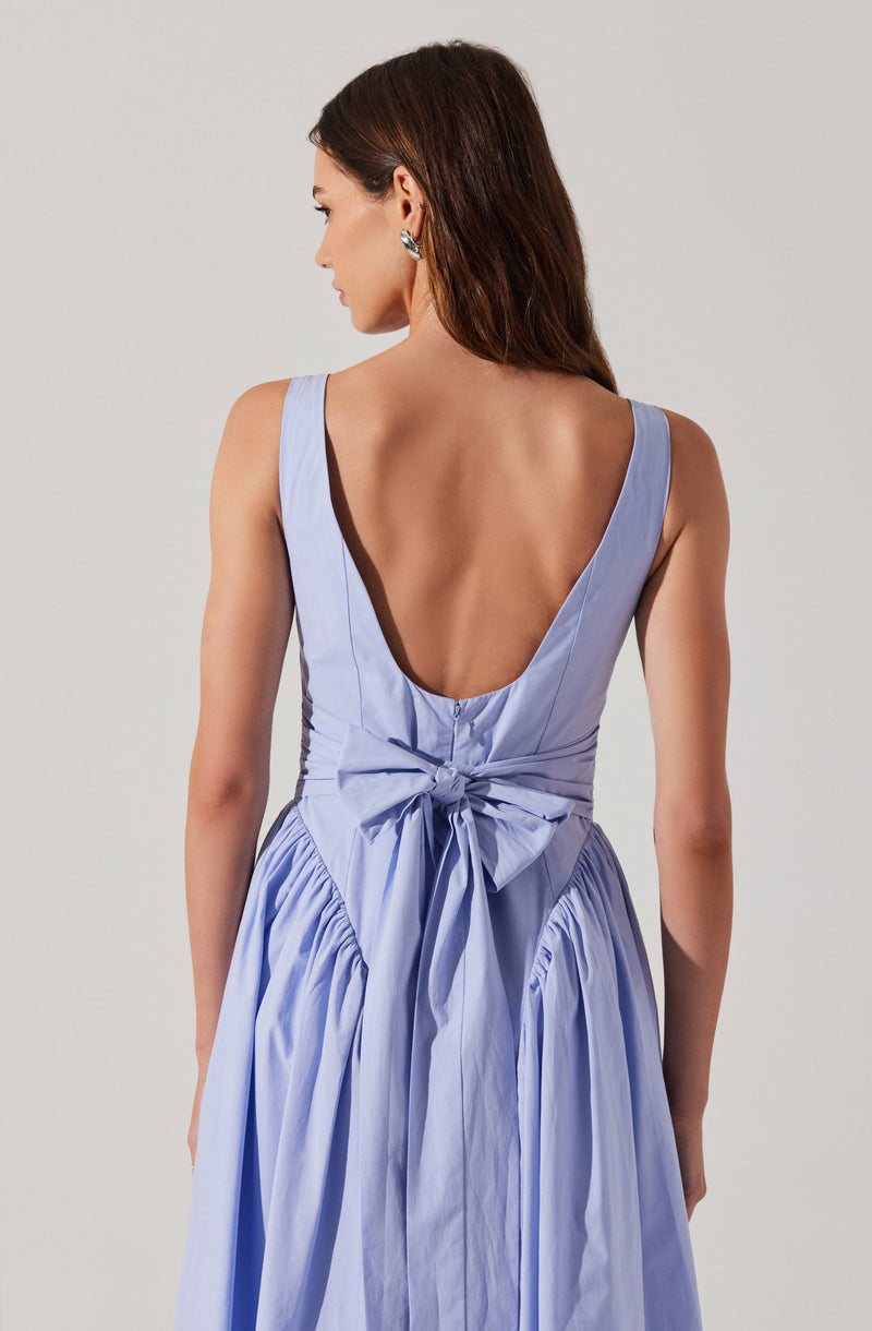 A woman is wearing a sleeveless, light blue dress with a deep V-back neckline. The dress features a fitted bodice with vertical seam details and a large bow tied at the waist. The skirt is gathered and has a full, flowing appearance with pleats.
