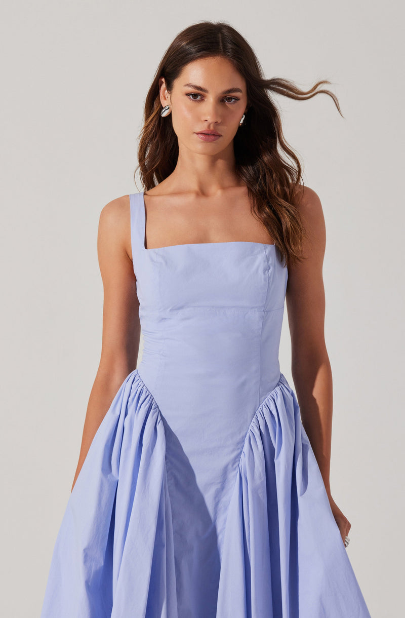 The woman is wearing a light purple, sleeveless dress with wide straps. The dress features a fitted bodice and a voluminous skirt with gathered fabric on the sides.