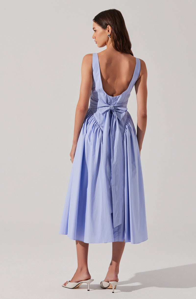 A woman is wearing a sleeveless, light blue dress with a deep V-back and a large bow tied at the back waist. The dress has a gathered, full skirt that reaches mid-calf length. She is also wearing white heeled shoes with a small bow detail.