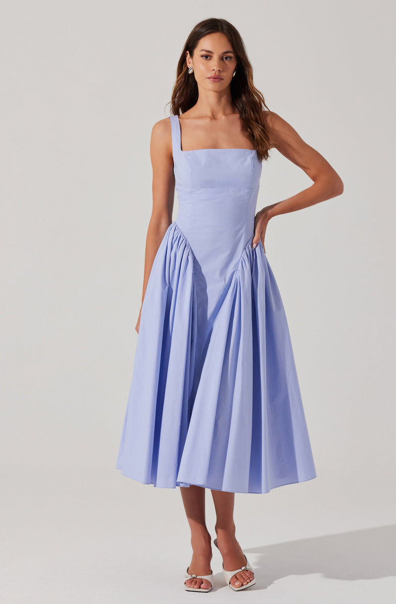 A woman is wearing a sleeveless, light blue dress with wide shoulder straps. The bodice is fitted, and the skirt is gathered with pleats, creating a voluminous, A-line silhouette that falls to mid-calf length. She is also wearing white open-toe heels.
