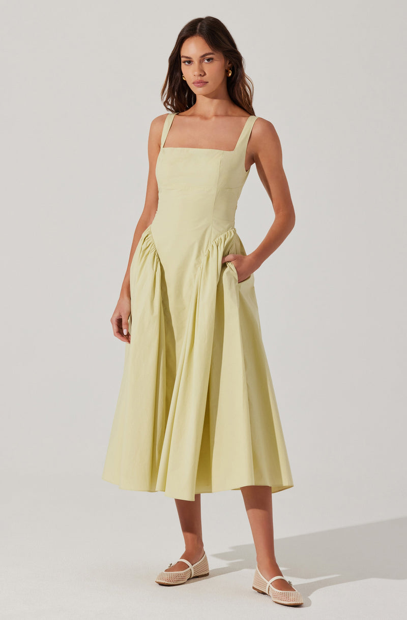 A woman is wearing a sleeveless, pale yellow dress with wide straps. The dress features a fitted bodice with a squared neckline and a full, gathered skirt that reaches mid-calf. She is also wearing light, woven flats.