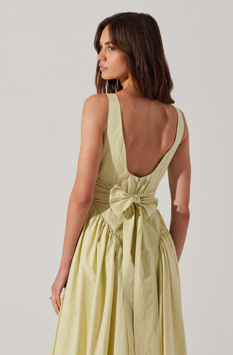 A woman is wearing a sleeveless, light yellow dress with a deep scoop back. The dress features a fitted bodice with a large, tied bow at the waist and a full, gathered skirt.