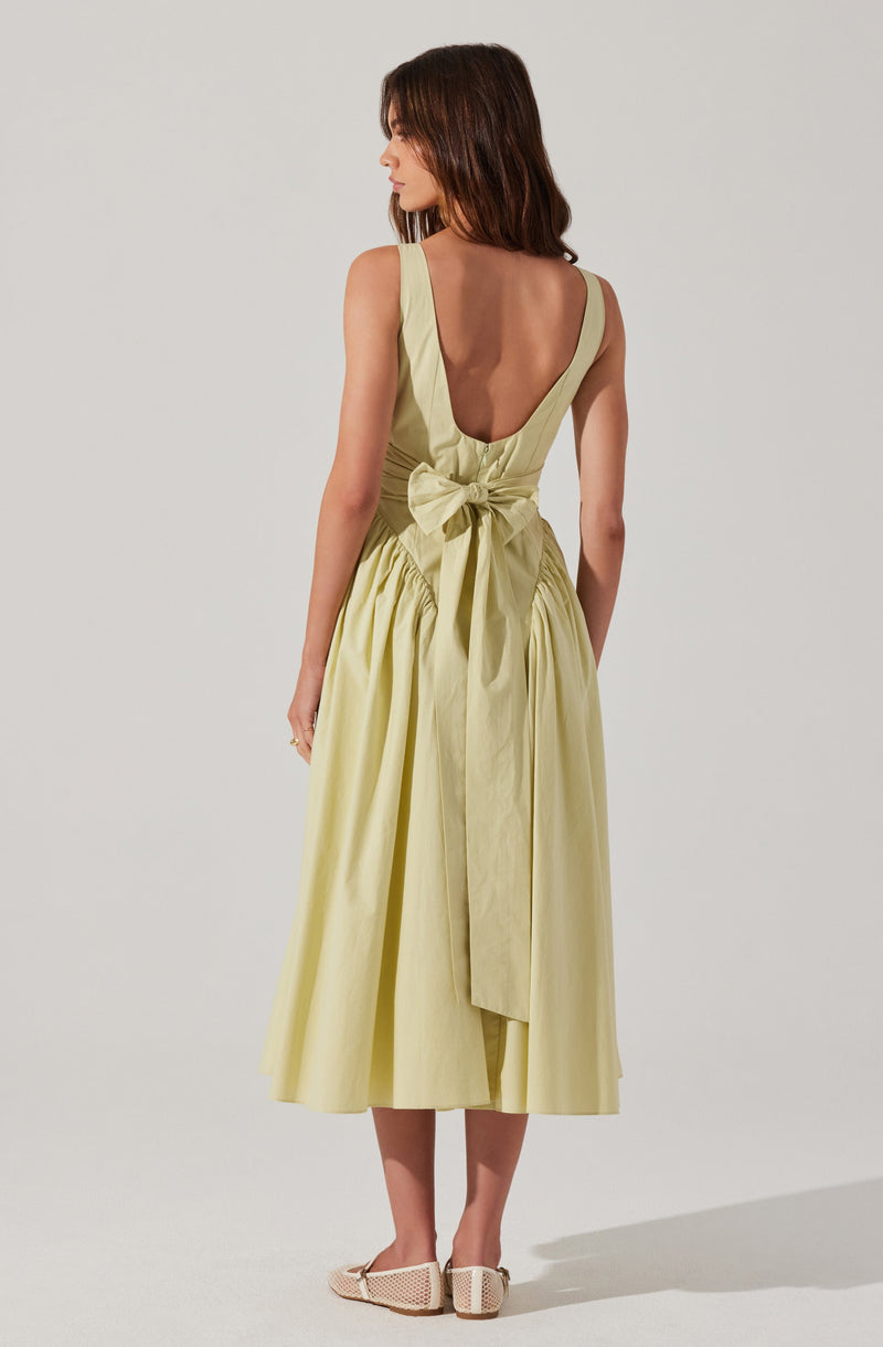 A woman is wearing a sleeveless, light yellow dress with a deep V-back. The dress features wide straps, a fitted waist with a large bow tied at the back, and a full, gathered skirt that falls below the knees. She is also wearing beige woven heeled shoes.