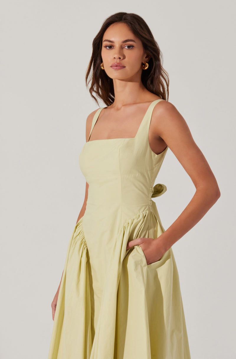 A woman is wearing a sleeveless, pale yellow dress with wide straps and a fitted bodice. The dress features a gathered skirt with pleats and a cut-out detail at the waist, tied with a bow at the back. She is accessorized with gold hoop earrings.
