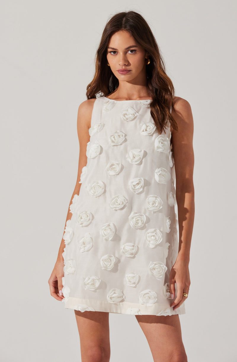 A woman is wearing a sleeveless, knee-length white dress decorated with three-dimensional fabric roses sewn throughout. The dress has a round neckline and a loose, straight fit.