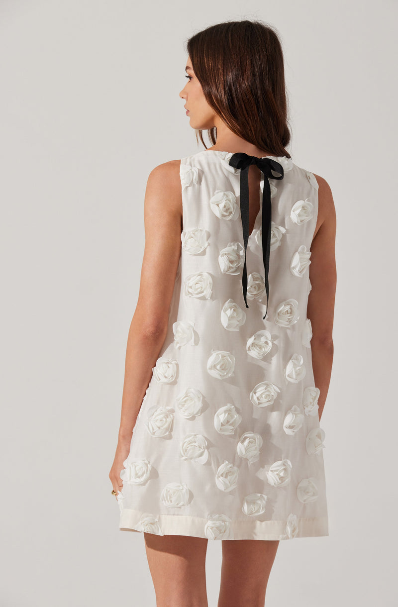 A woman is wearing a sleeveless, knee-length white dress decorated with 3D white rose embellishments scattered throughout the fabric. The dress features a round neckline with a black ribbon tied into a bow at the back neck. The fabric appears lightweight and semi-sheer.
