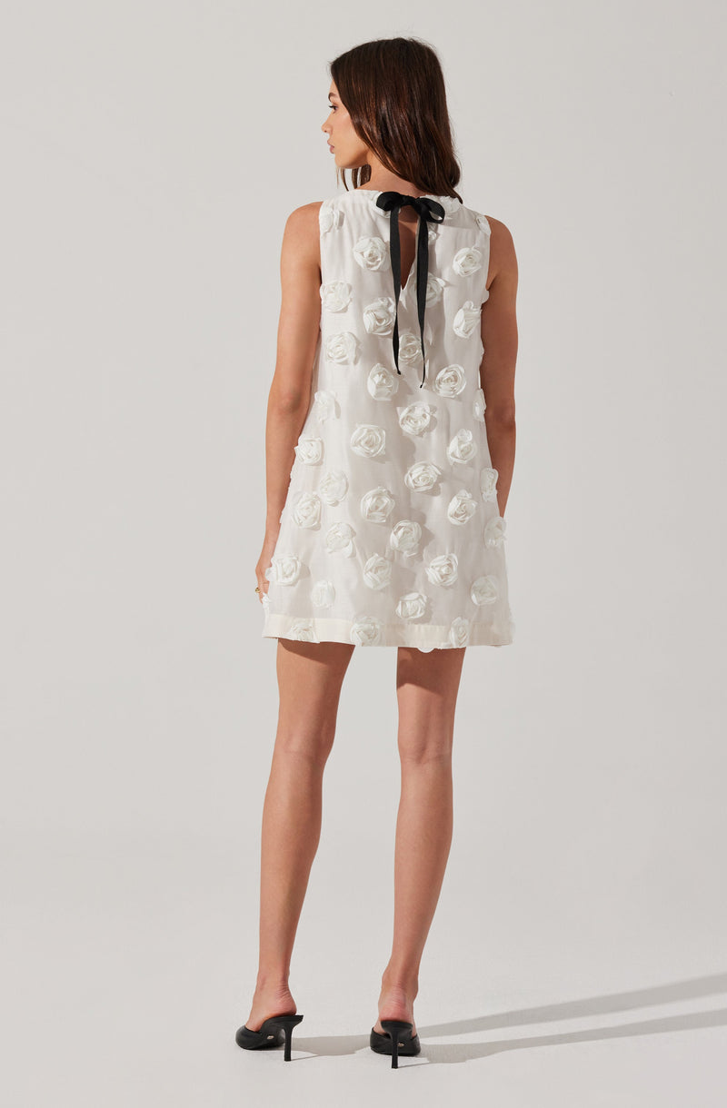 The dress is sleeveless and white, decorated with textured, three-dimensional floral patterns made of fabric roses throughout. It features a black bow at the neckline, with the tie hanging down the back. The dress has a loose, A-line silhouette and ends above the knees.