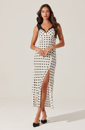 A woman is wearing a sleeveless, ankle-length dress with a white background and black polka dots. The dress features black lace trim along the V-neckline and shoulder straps. It has a high slit on one side, revealing her leg. She is also wearing black pointed-toe shoes with bows.