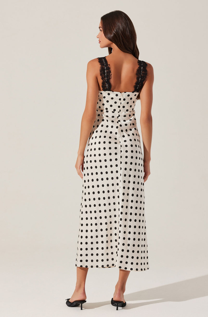 A woman is wearing a sleeveless jumpsuit with a fitted bodice and wide-leg pants. The jumpsuit features a white background with black polka dots and has black lace straps. She is also wearing black high-heeled sandals.