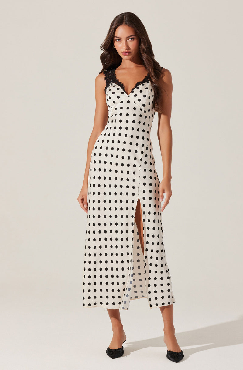 A woman is wearing a sleeveless, full-length maxi dress with a white background and black polka dots. The dress features black lace trim along the V-neckline and shoulder straps. It has a front slit that extends from the hem to just above the knee. She is also wearing black pointed-toe heels with a bow detail at the front.