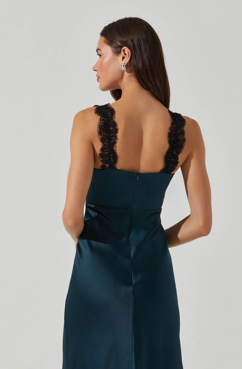 A woman is wearing a sleeveless, dark teal satin dress with black lace straps. The dress has a fitted bodice and a zipper closure at the back. The lace straps feature a floral pattern. The woman has long, dark hair and is accessorized with earrings.