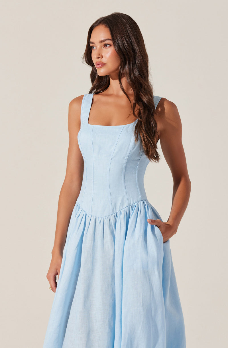 A woman is wearing a sleeveless, light blue dress with wide shoulder straps, a fitted bodice with vertical seams, and a gathered skirt that falls to a midi length. The dress has a square neckline and appears to be made of a lightweight, woven fabric.