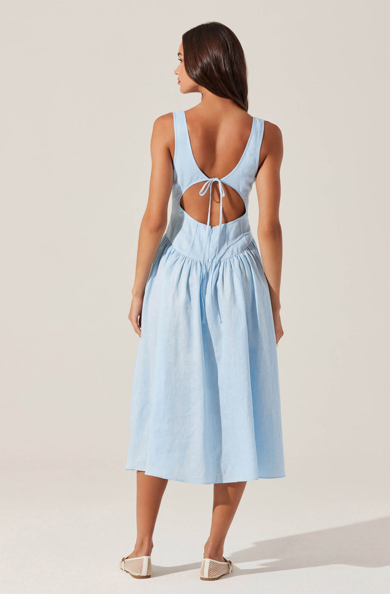 A woman is wearing a light blue sleeveless dress with a fitted bodice and wide straps. The dress features a large circular cutout at the back, framed by the straps, and is tied with a bow at the top of the cutout. The skirt of the dress is gathered at the waist, flowing down to a mid-calf length. She is also wearing light-colored pointed-toe shoes with a textured surface.