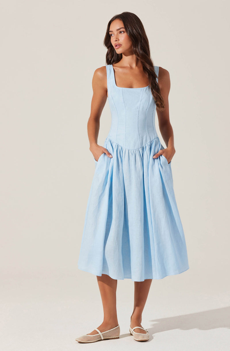 A woman is wearing a light blue sleeveless dress with a fitted bodice featuring vertical seam details and a square neckline. The dress has a full, gathered skirt that falls to mid-calf length and includes pockets, which the woman is resting her hands in. She is also wearing beige woven flat shoes.