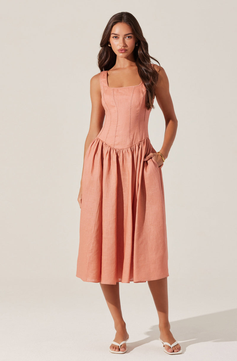 A woman is wearing a peach-colored sleeveless dress with a fitted, corset-style bodice that features vertical seam details. The dress has a square neckline and a full, gathered skirt that falls below the knees. She is also wearing white open-toe sandals with a low heel.