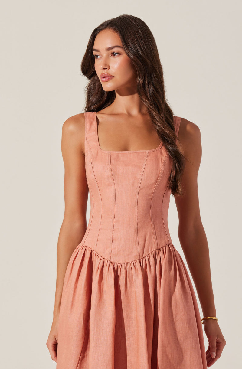 A woman is wearing a sleeveless, peach-colored dress with a fitted bodice featuring vertical seam details and a scoop neckline. The skirt of the dress is gathered at the waist, creating a full, flowing appearance.