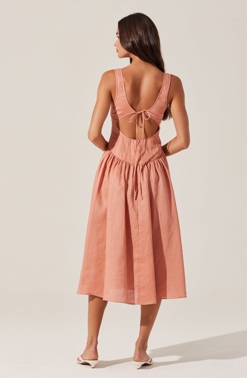 The woman is wearing a sleeveless, peach-colored dress with a fitted bodice and wide shoulder straps. The back of the dress features a large oval cutout with a tie closure at the top and a drawstring detail at the waist. The skirt of the dress is gathered and has a midi length, falling below the knees.