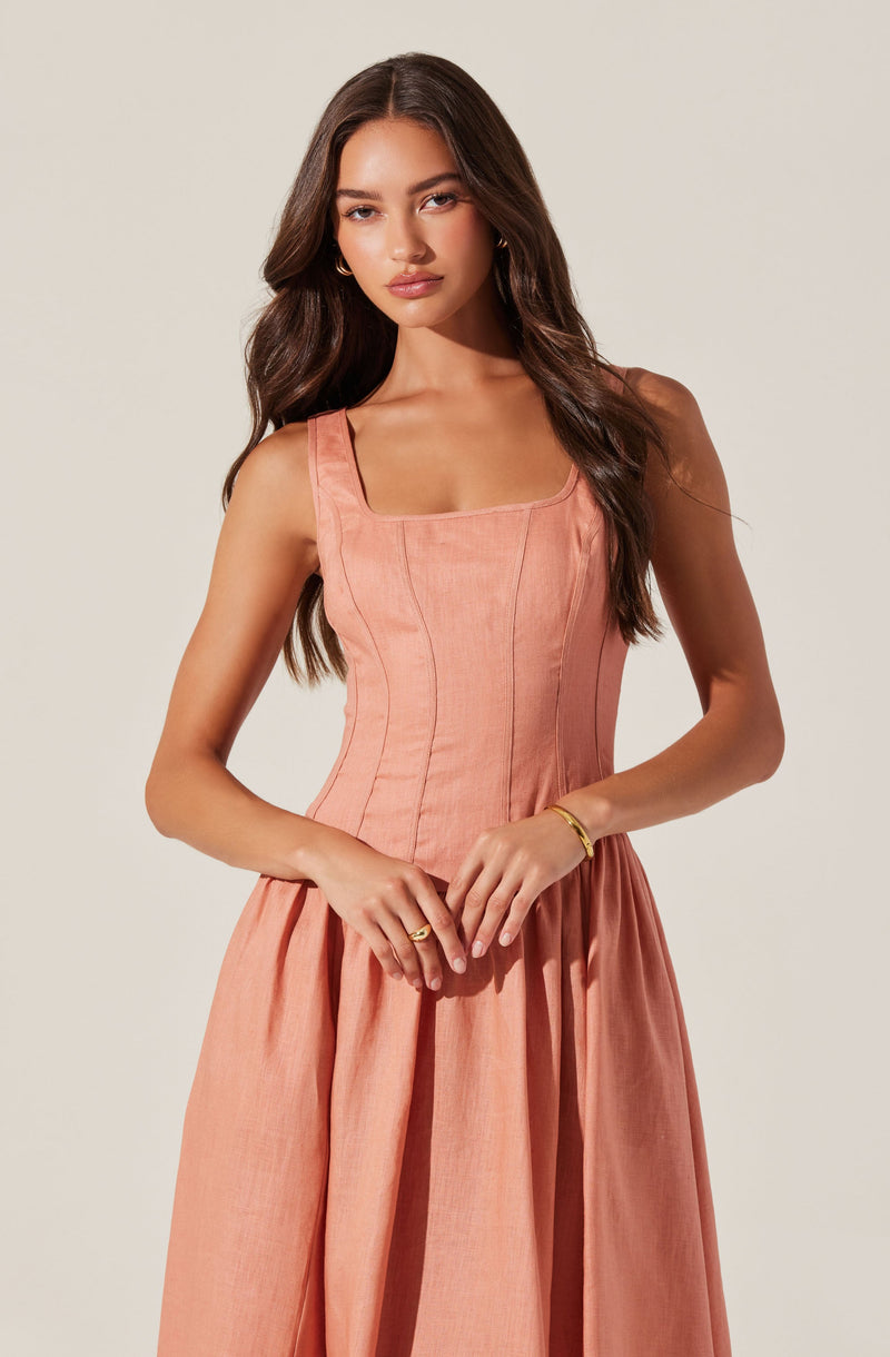 A woman is wearing a sleeveless, peach-colored dress with a fitted bodice featuring vertical seam lines and a square neckline. The skirt is full and gathered, creating a draped, flowing effect.