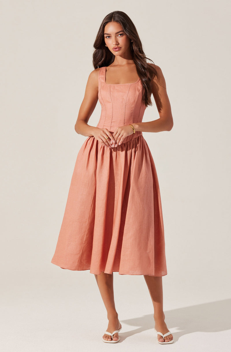 A woman is wearing a sleeveless peach-colored dress with a fitted bodice that has vertical stitching details and a square neckline. The dress features a full, gathered skirt that falls below the knees. She is also wearing white open-toe sandals with a small heel and a simple bracelet on her wrist.