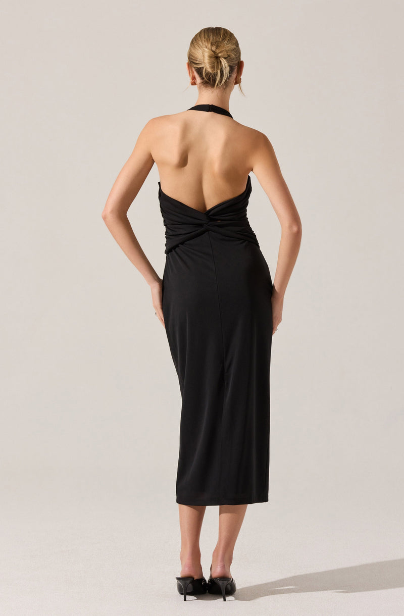 A woman wearing a black halter neck dress with a ruched bodice that wraps around the waist. The dress is sleeveless, backless, and has a midi length that falls below the knees. She is also wearing black high heels.