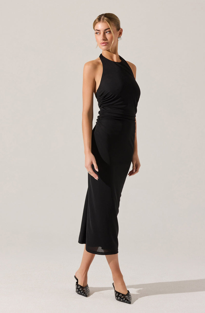 A woman is wearing a sleeveless black halter-neck dress that is fitted around the waist with ruched detailing and a long, flowing skirt that reaches mid-calf. She is also wearing black high-heeled shoes adorned with metallic eyelet details.