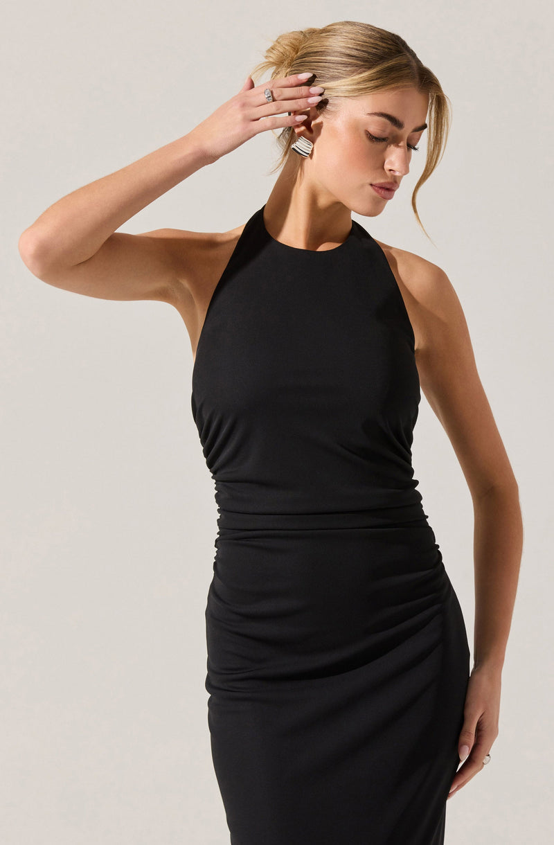 A woman is wearing a sleeveless, black halter neck dress that is fitted and gathered around the waist and hips. She has her hair styled in an updo and is accessorized with large rectangular earrings and rings on her fingers.