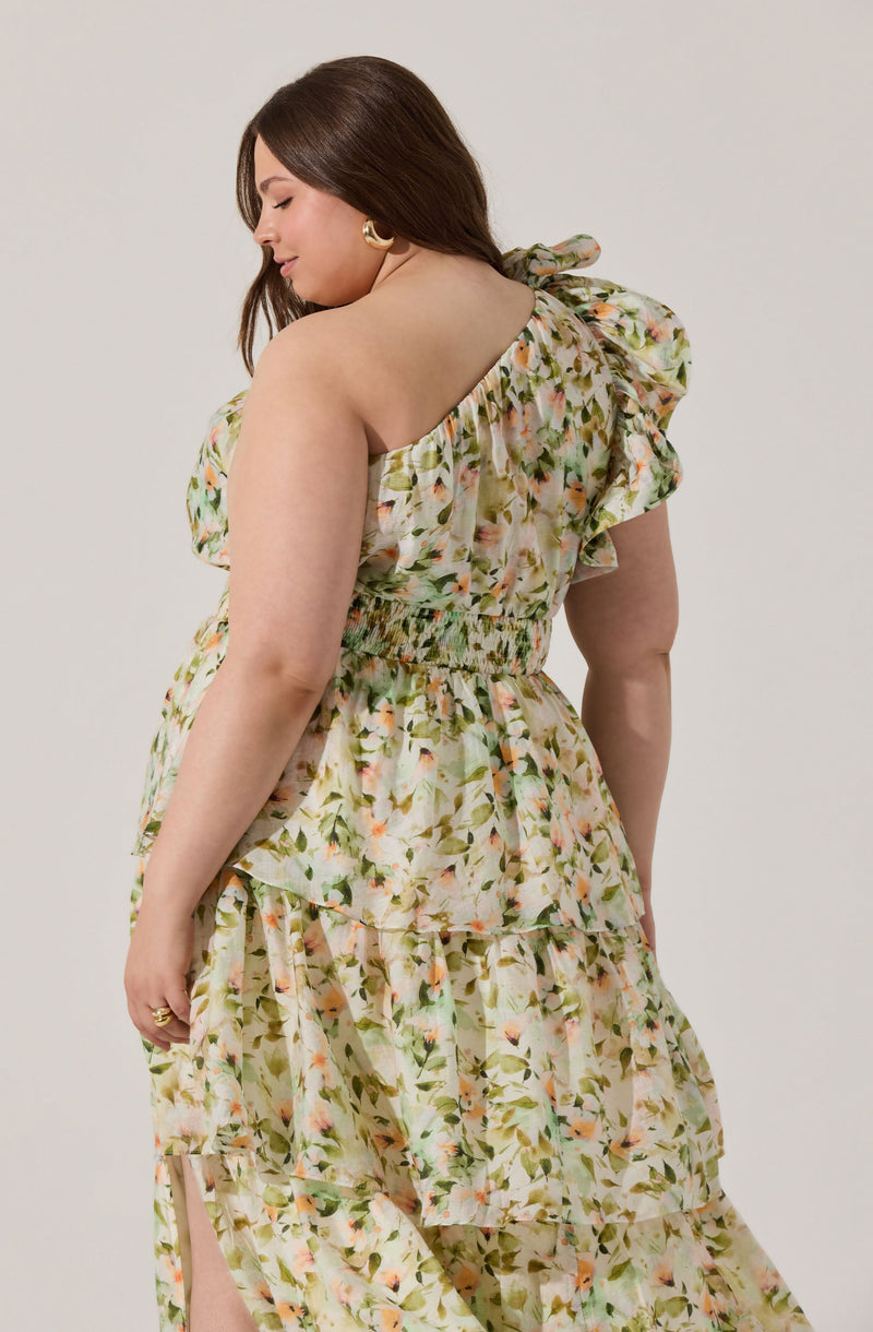 A woman is wearing a sleeveless, floral-patterned dress with ruffled details on the shoulder and a fitted, smocked waist. The fabric features a light, pastel-colored print with various shades of green, peach, and pink.