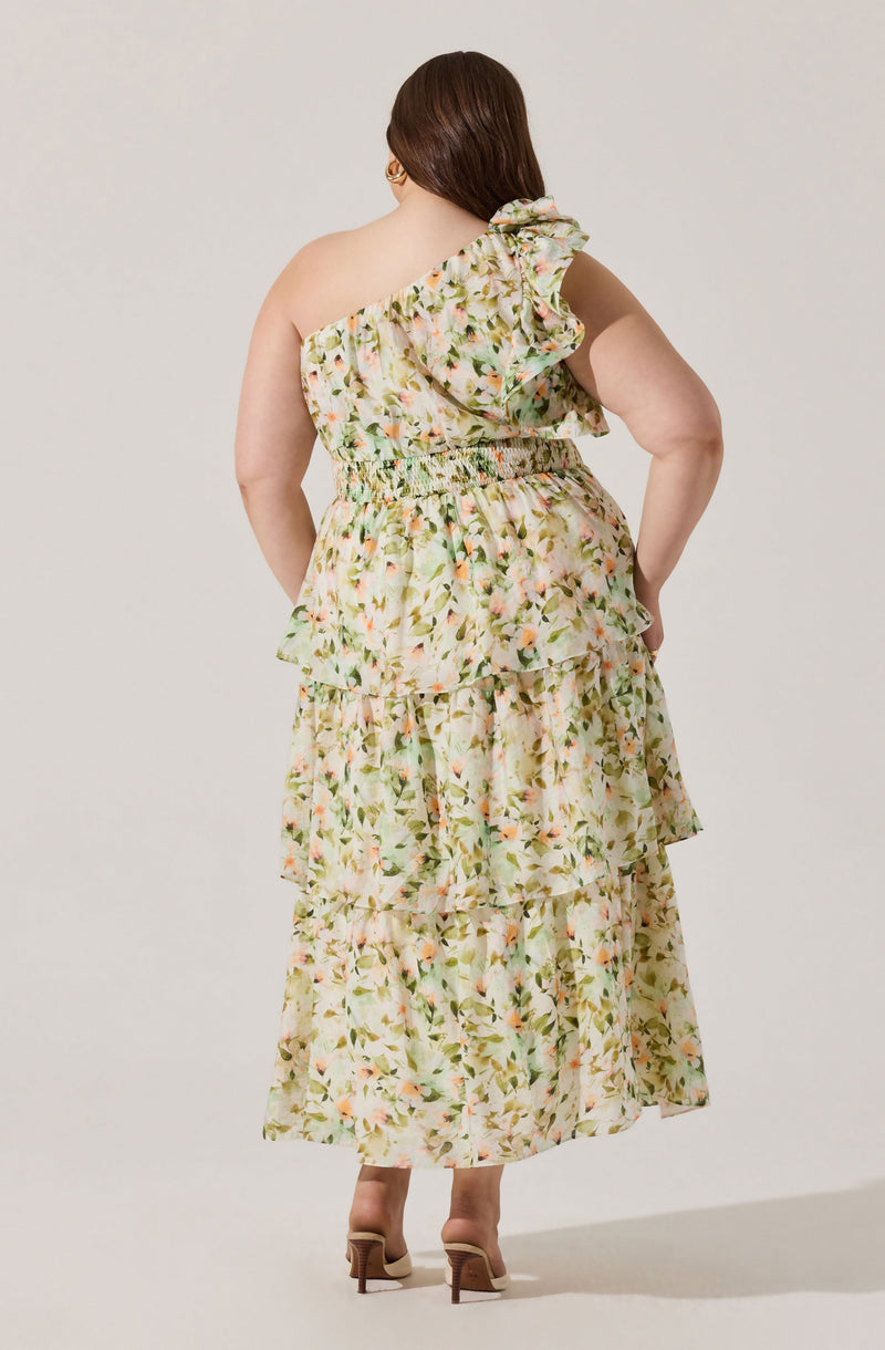 A woman is wearing a long, flowy dress with a floral print in soft pastel colors. The dress features a one-shoulder design with a large ruffle detail on the shoulder and around the neckline. The waist is gathered with elastic, creating a cinched effect, and the skirt is tiered with multiple ruffled layers, giving it a layered, voluminous look. The fabric has a light, airy appearance suitable for warm weather. She is also wearing beige high-heeled shoes.