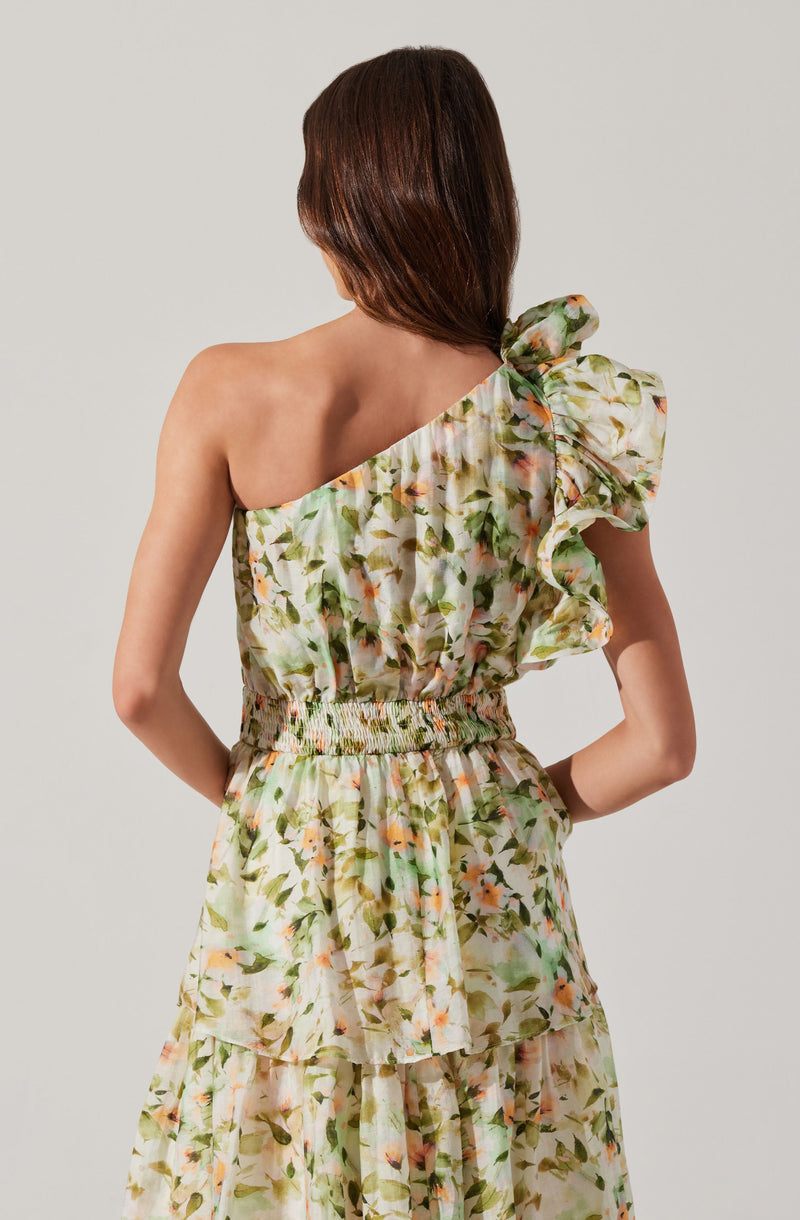 A woman is wearing a floral-patterned dress with a one-shoulder design. The dress has a ruffled sleeve on the right shoulder, a smocked waistband, and a layered, flowing skirt featuring the same floral pattern with soft, pastel-colored flowers and green leaves.