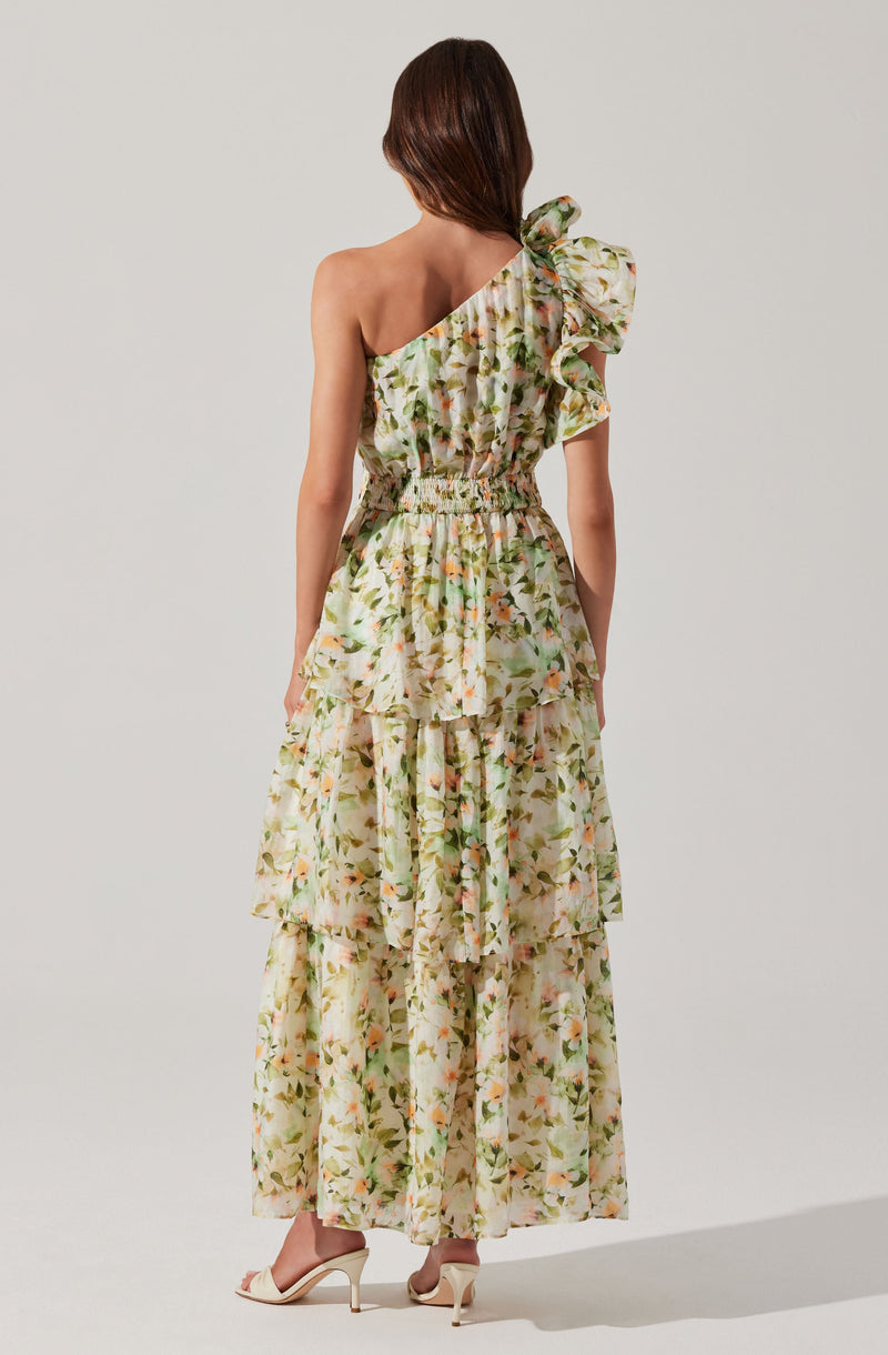 A woman is wearing a long, flowing floral dress with a one-shoulder design. The dress features a ruffled strap on the left shoulder and an elastic waistband. The fabric has a light, airy appearance with a pattern of soft-colored flowers and green leaves throughout.