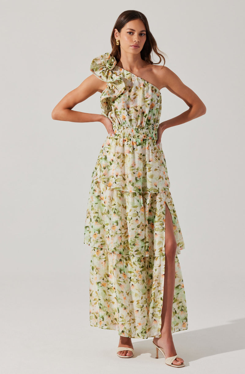 A woman is wearing a long, flowy dress with a floral pattern in shades of green, beige, and pink. The dress features an asymmetrical, one-shoulder design with a large ruffle on the shoulder. It has an elastic waistband and a high slit on the left side that reveals her leg.