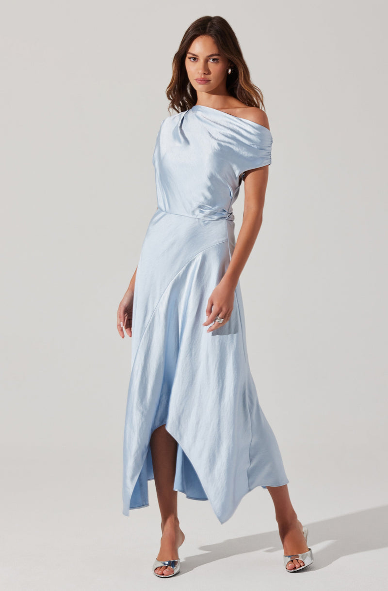 A woman is wearing a light blue satin dress with a draped, off-the-shoulder neckline. The dress has a fitted waist and a flowing, asymmetrical hemline that is longer in the back and shorter in the front. She is also wearing open-toe high heels with a metallic finish and has earrings and rings as accessories.