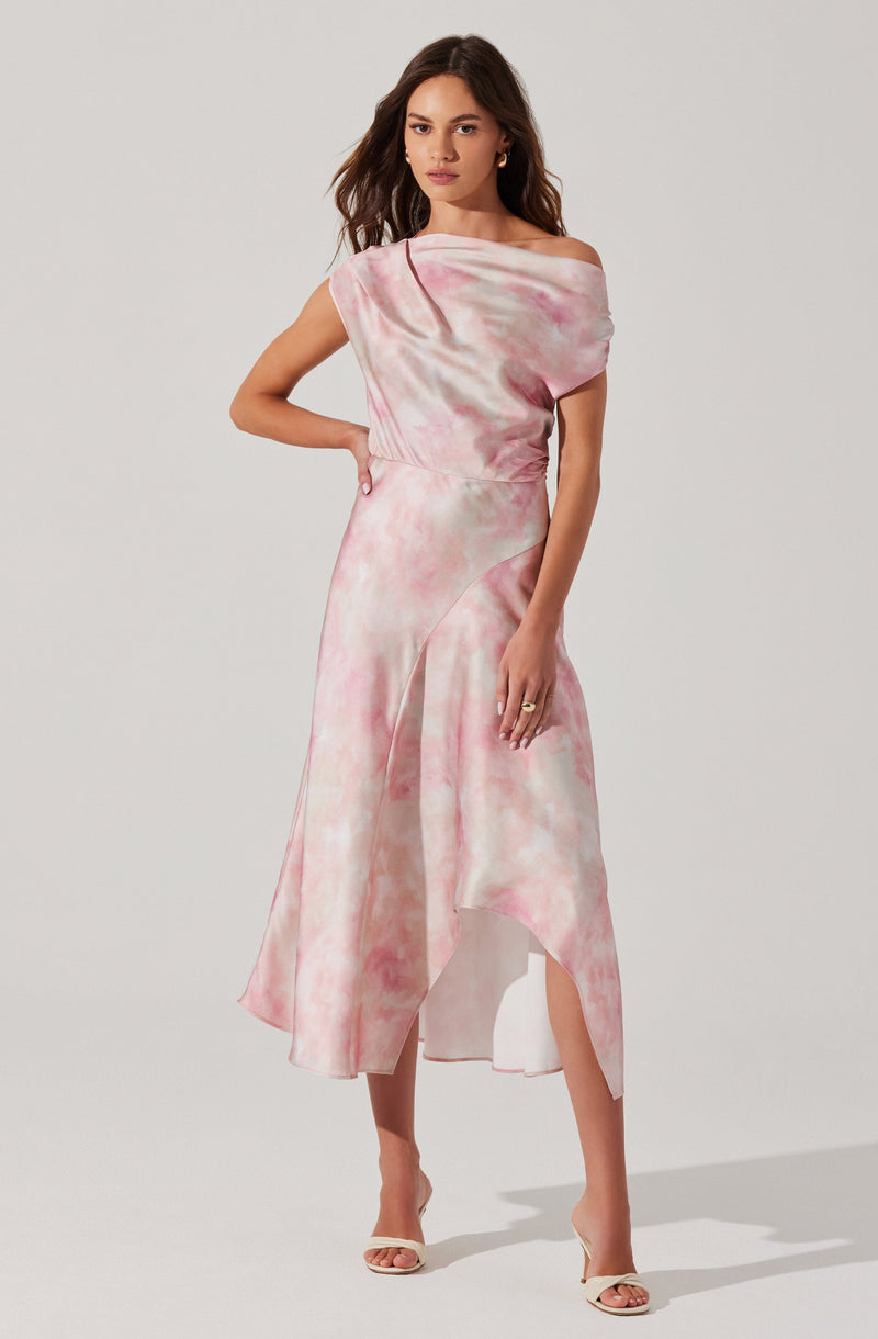 A woman is wearing a flowing, elegant dress with an off-the-shoulder neckline. The dress features a soft, pastel pink and cream tie-dye pattern and has a high-low hemline with slits on the sides, revealing her legs. She is also wearing open-toe heels in a neutral color.