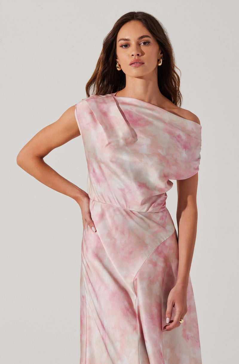 A woman is wearing a sleeveless, off-the-shoulder dress with a pink and white tie-dye pattern. The fabric appears silky and drapes smoothly over her body. She has wavy brown hair and is accessorizing with gold hoop earrings.