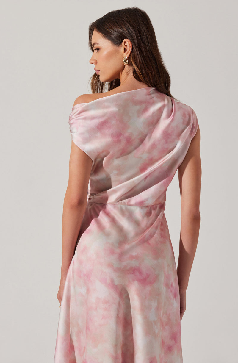 The dress is a sleeveless, flowing gown with a one-shoulder design. It features a soft, watercolor-like pattern in shades of pink, cream, and light gray, creating a delicate and elegant appearance. The fabric appears to be smooth and silky, with gentle draping and subtle gathers.