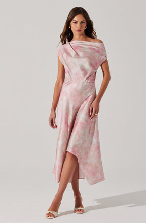 A woman is wearing a sleeveless, off-the-shoulder dress with a pink and white tie-dye pattern. The dress has a fitted waist and a flowing, asymmetrical hemline that is longer in the back and higher in the front, revealing her legs. She is also wearing open-toe heels and gold hoop earrings.
