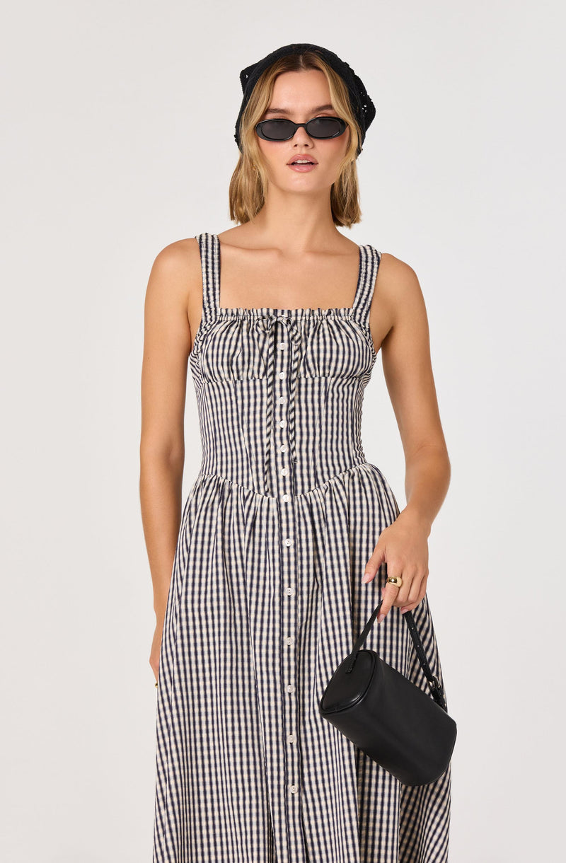 Young woman wearing a black cap, sunglasses, and a sleeveless, checkered dress with vertical stripes.