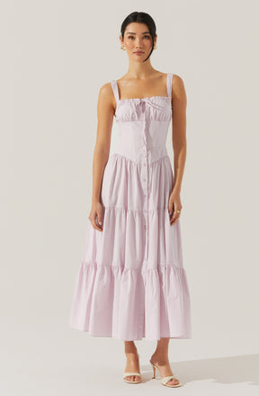 A woman is wearing a light pink, sleeveless, tiered maxi dress with a square neckline and adjustable straps. The dress features a fitted bodice with a front button closure and has gathered tiers from the waist down, creating a flowing silhouette. She is also wearing beige high-heeled sandals.