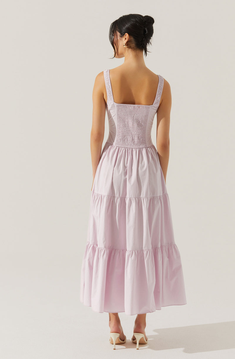 A woman is wearing a sleeveless, light pink maxi dress with wide shoulder straps. The bodice is fitted with gathered or smocked detail, and the skirt is flowing with multiple tiered sections. She is also wearing light-colored high-heeled shoes.