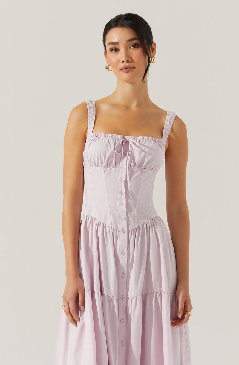 A woman is wearing a sleeveless, light pink sundress with wide straps that have lace details. The dress features a fitted bodice with gathered fabric at the top, a row of buttons running down the front, and a gathered, tiered skirt.