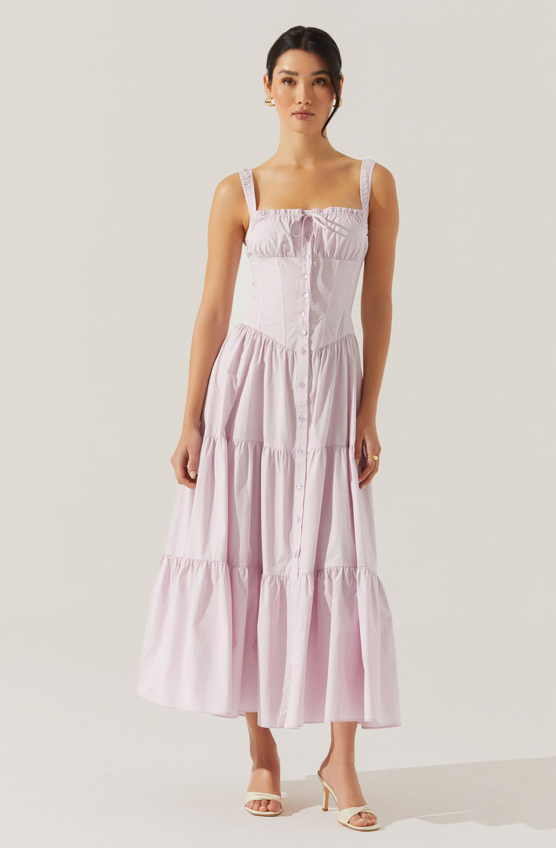 A woman is wearing a sleeveless, light pink, tiered maxi dress with wide shoulder straps and a front button-down design. The dress has a gathered bodice with a small bow at the center of the neckline. She is also wearing open-toed, high-heeled sandals.
