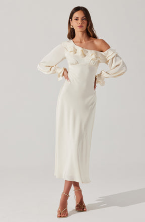 A woman is wearing a long, cream-colored dress with an off-the-shoulder neckline. The dress features ruffled details around the neckline and on the long, puffed sleeves that gather at the wrists. She has paired the dress with strappy high-heeled sandals.