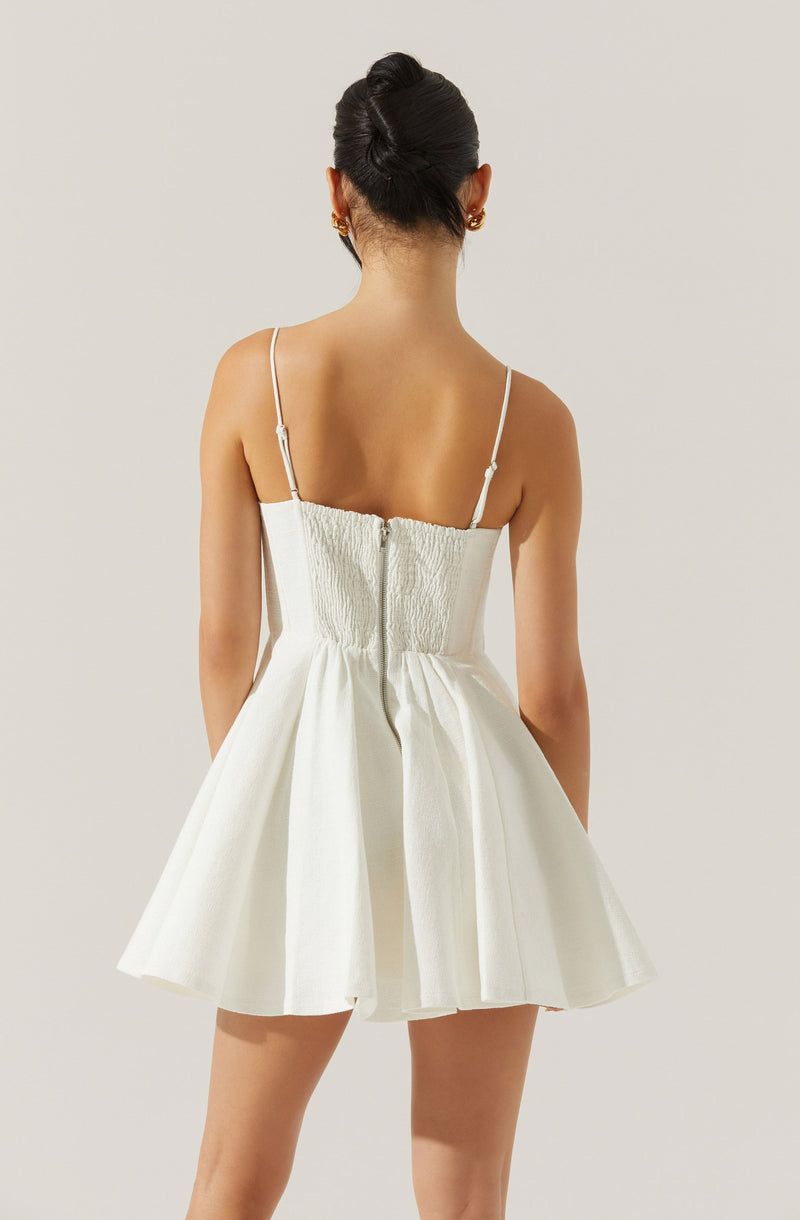 A woman is wearing a white dress with thin spaghetti straps. The top part of the dress features a textured, ruched fabric, and the skirt is pleated with a flared, A-line silhouette. The dress appears to be made of a lightweight, structured material.
